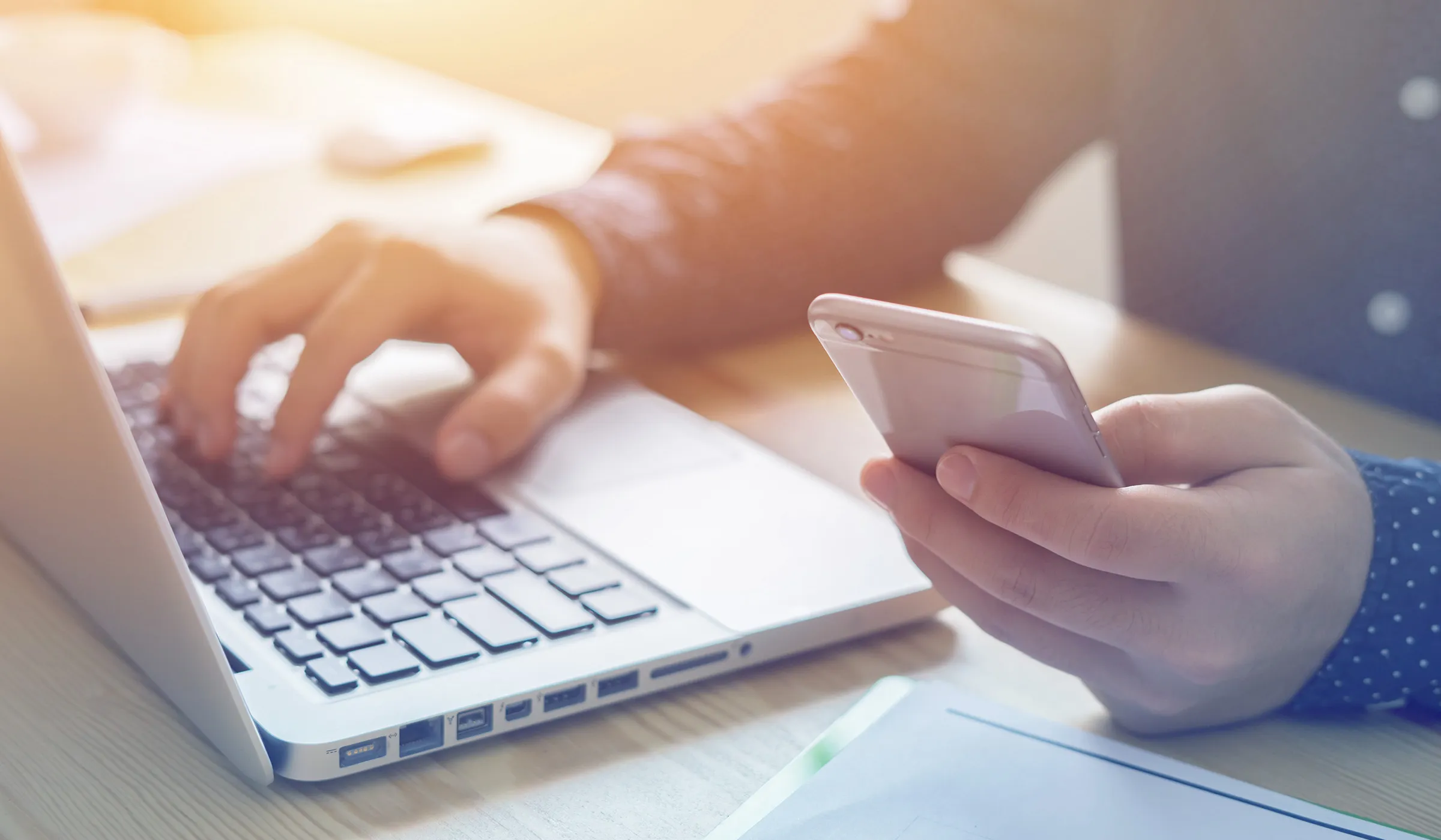  Eine Person sitzt vor einem Laptop, eine Hand ruht auf der Tastatur, während die andere ein Handy hält. Die Szene vermittelt Multitasking und effiziente Nutzung von Technologien für verschiedene Aufgaben.