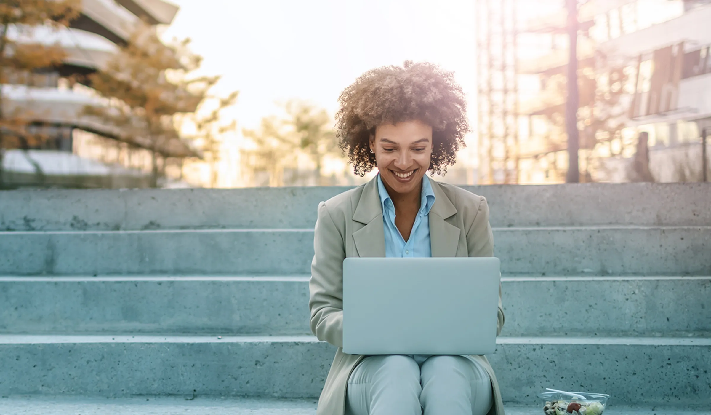 Eine Frau sitzt entspannt auf einer Treppe mit einem Laptop auf ihrem Schoß. Sie ist in eine Arbeit vertieft oder genießt ihre Umgebung. Der Laptop zeigt Professionalität und Flexibilität im Arbeitsstil.
