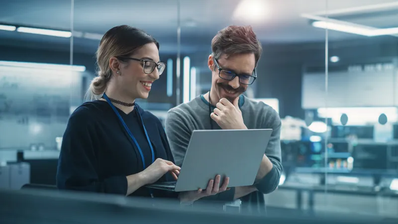 Ein Brainguards Mitarbeiter und eine Brainguards Mitarbeiterin stehen lachend im Großraumbüro und schauen auf den Laptop, den die Frau in den Händen hält. Die Szene strahlt eine positive und kooperative Arbeitsatmosphäre aus, in der gemeinsames Engagement und Freude am Arbeitsplatz sichtbar werden.