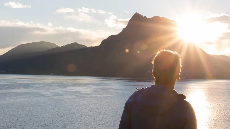 Eine Person steht vor einer beeindruckenden Berglandschaft mit einem See im Vordergrund. Hinter den majestätischen Bergen erscheint die Sonne, die ihr warmes Licht über die Szenerie wirft. Ein atemberaubender Anblick, der Ruhe und Naturverbundenheit vermittelt.