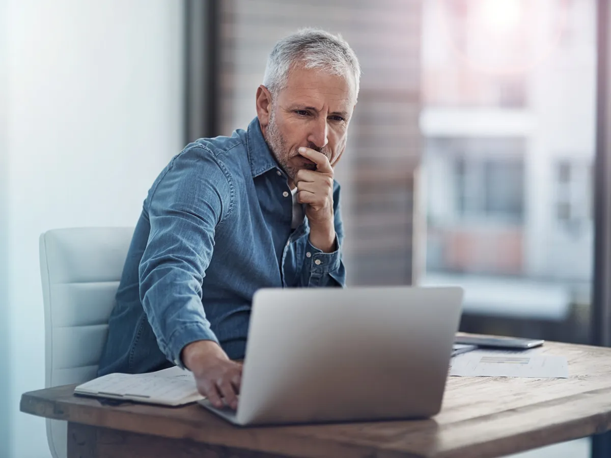 Ein Mann sitzt am Schreibtisch vor seinem Laptop, wobei seine Hand auf der Tastatur liegt. Sein Gesichtsausdruck zeigt Skepsis, während er den Bildschirm aufmerksam betrachtet. Die Szene deutet auf eine intensive Überlegungen hin.