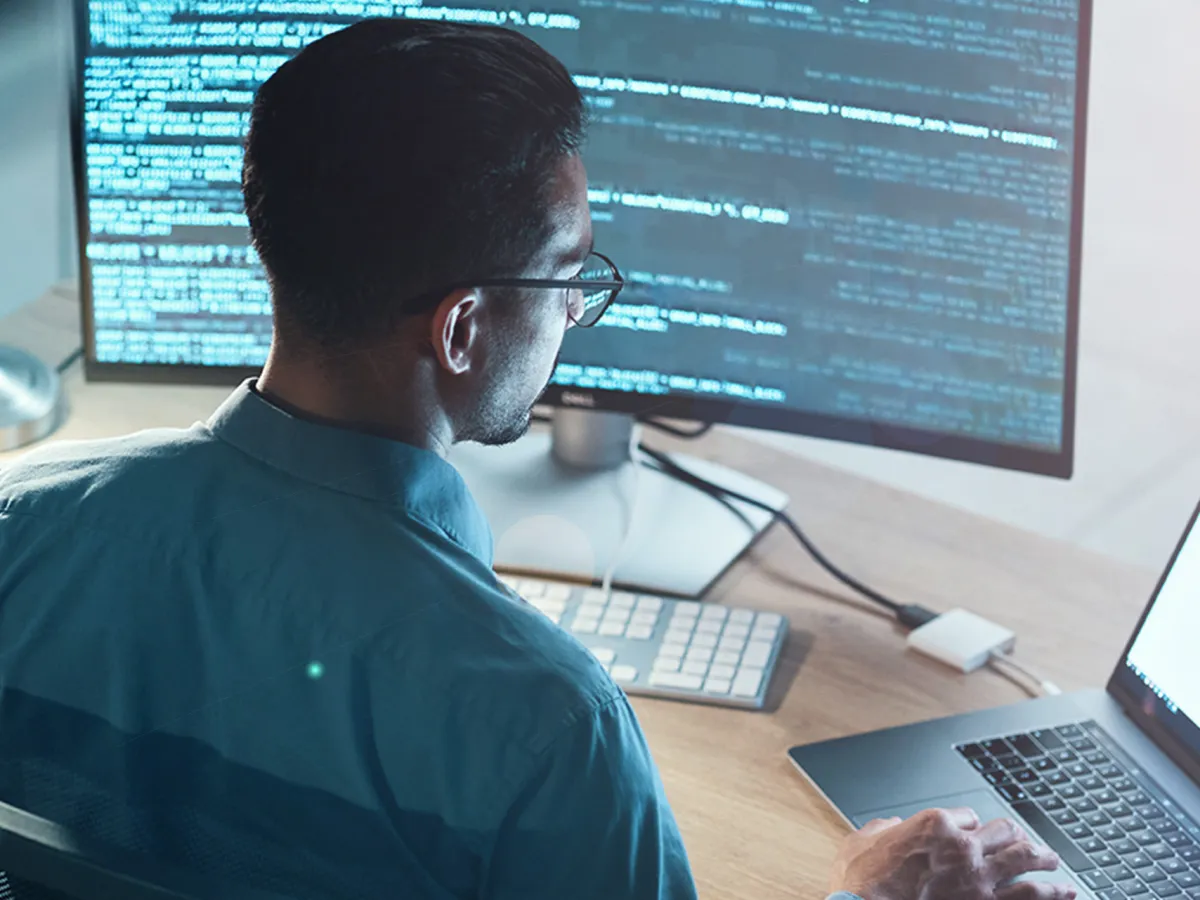 Ein Mann sitzt vor einem Monitor mit Quellcode und betrachtet ihn angestrengt. Sein fokussierter Blick zeigt, dass er in die Details des Codes vertieft ist.